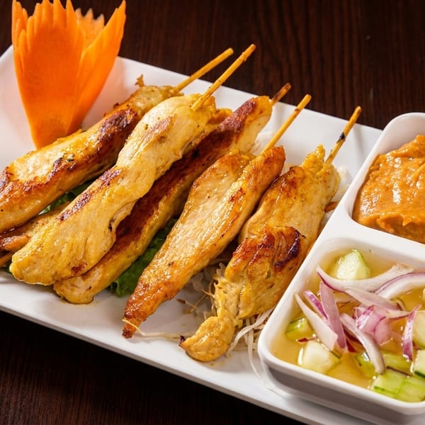 Chicken satay skewers with peanut sauce and cucumber salad on a white rectangular plate.