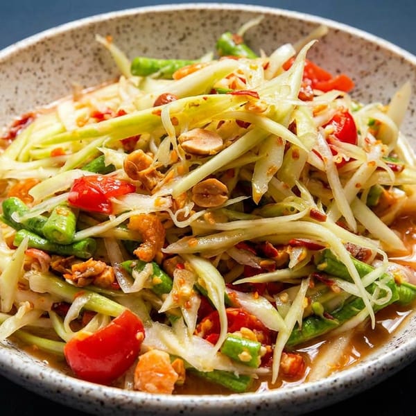 Close-up of a vibrant Thai papaya salad with peanuts, tomatoes, and green beans in a speckled bowl.