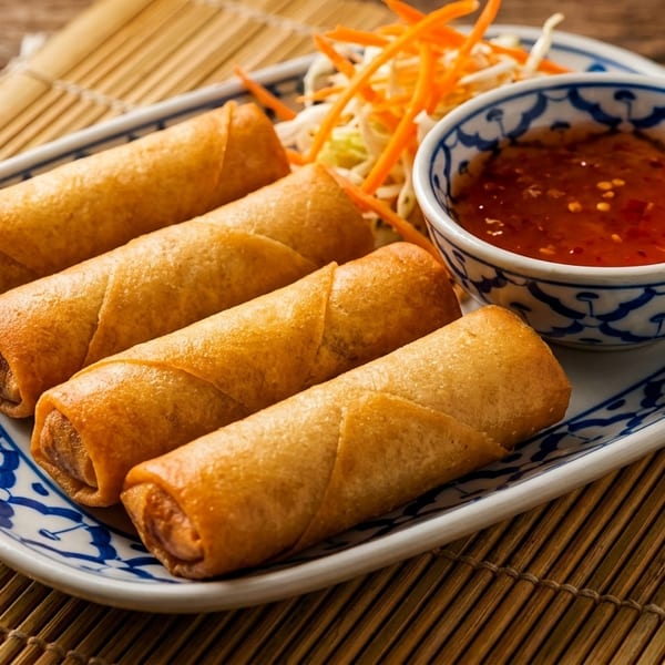 Four golden-brown spring rolls with sweet chili sauce and shredded vegetables on a blue and white plate.
