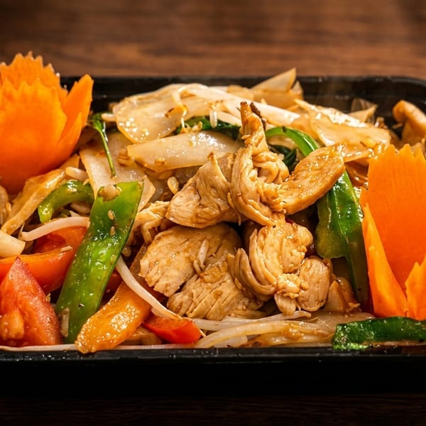 Close-up of a chicken stir-fry with noodles, vegetables, and decorative carrot garnishes in a black container.